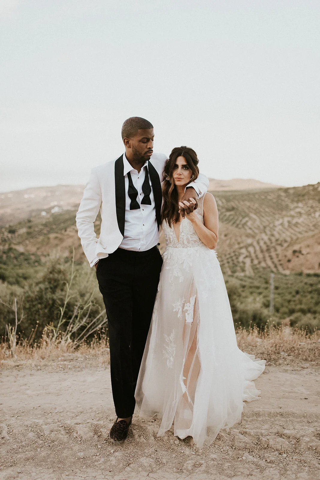 A bride and groom standing outdoors in a desert landscape, with the groom in a white tuxedo with a black bowtie and the bride in a lace wedding dress, holding hands and looking at each other.