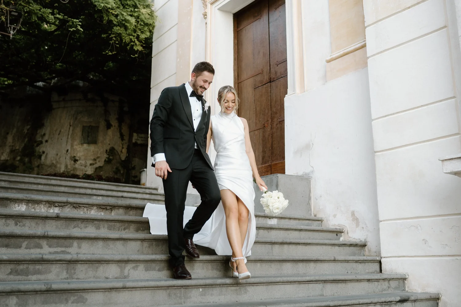 A newlywed couple descending outdoor stone steps after their wedding. The groom is wearing a black tuxedo with a bow tie, and the bride is in a white wedding dress holding a bouquet of white flowers. They are smiling and looking down as they walk tog