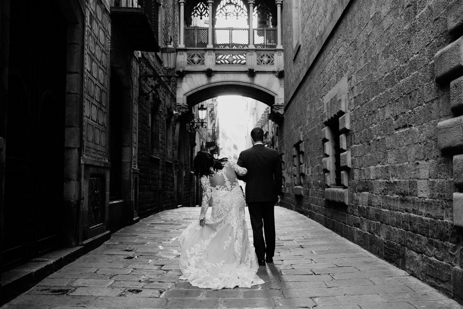 A bride and groom in wedding attire walking hand in hand through a narrow, historic alleyway with brick walls and an archway overhead, in black and white.