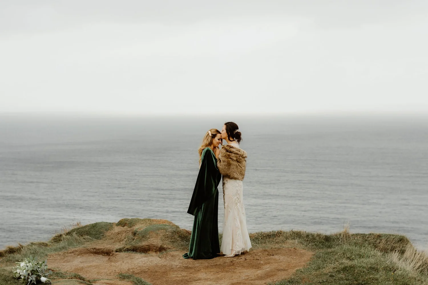 Two women dressed in formal attire embracing each other on a rocky cliff by the ocean, with one woman kissing the other's forehead.