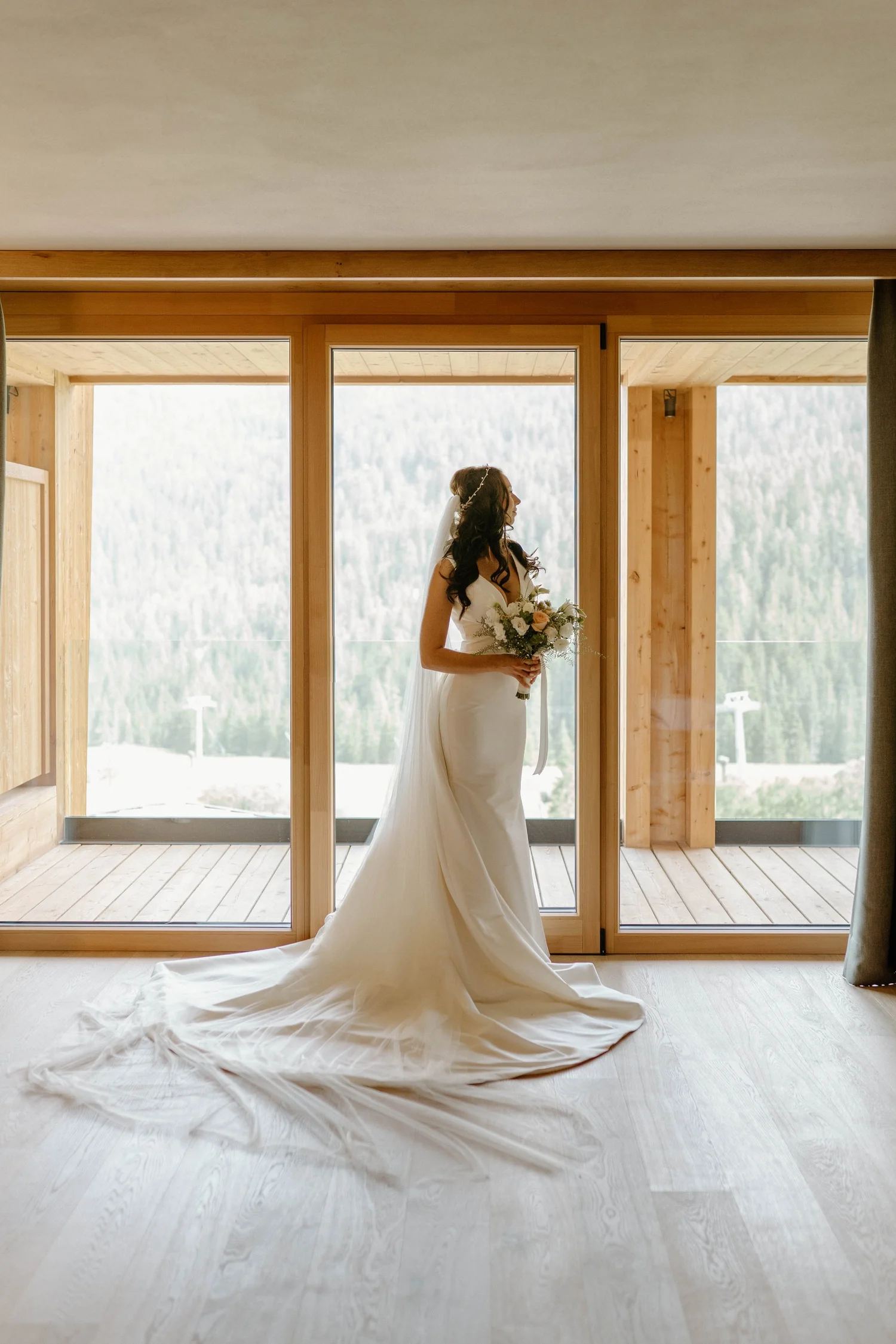 A bride in a white wedding dress holding a bouquet of flowers, standing indoors near a large glass sliding door with a scenic mountain view in the background.