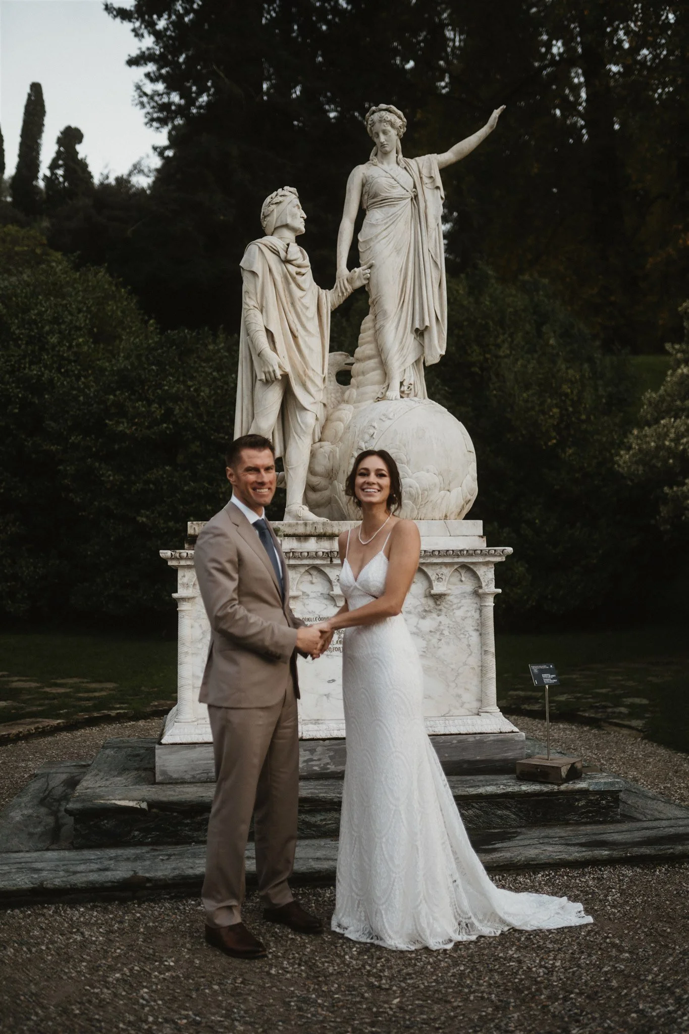 A newlywed couple in formal wedding attire standing outdoors in front of a classical marble statue. The couple is holding hands and smiling at the camera.