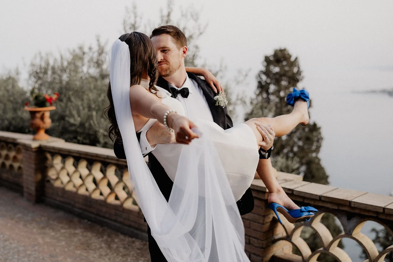 A groom in a tuxedo holding a bride in a white dress with a veil and blue high heels, outdoors with trees and a decorative railing in the background.