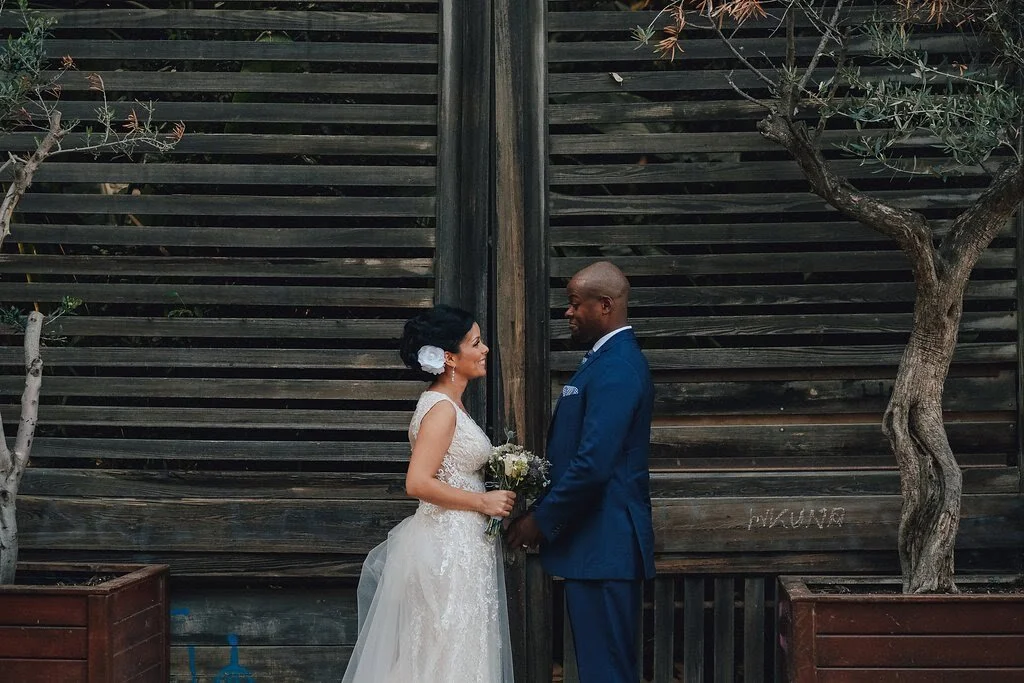 A bride and groom holding hands and facing each other outdoors, with a wooden fence in the background and trees on either side.