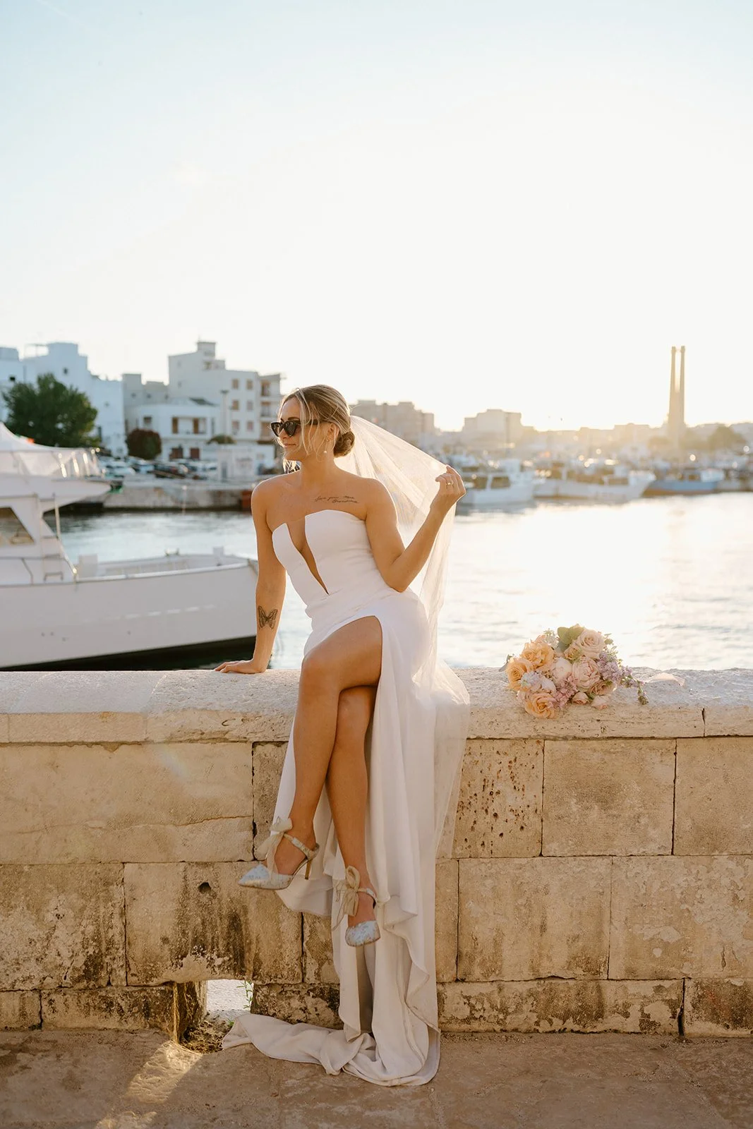 A woman in a white wedding dress sitting on a stone ledge by the water at sunset, with boats and buildings in the background and a bouquet of flowers beside her.