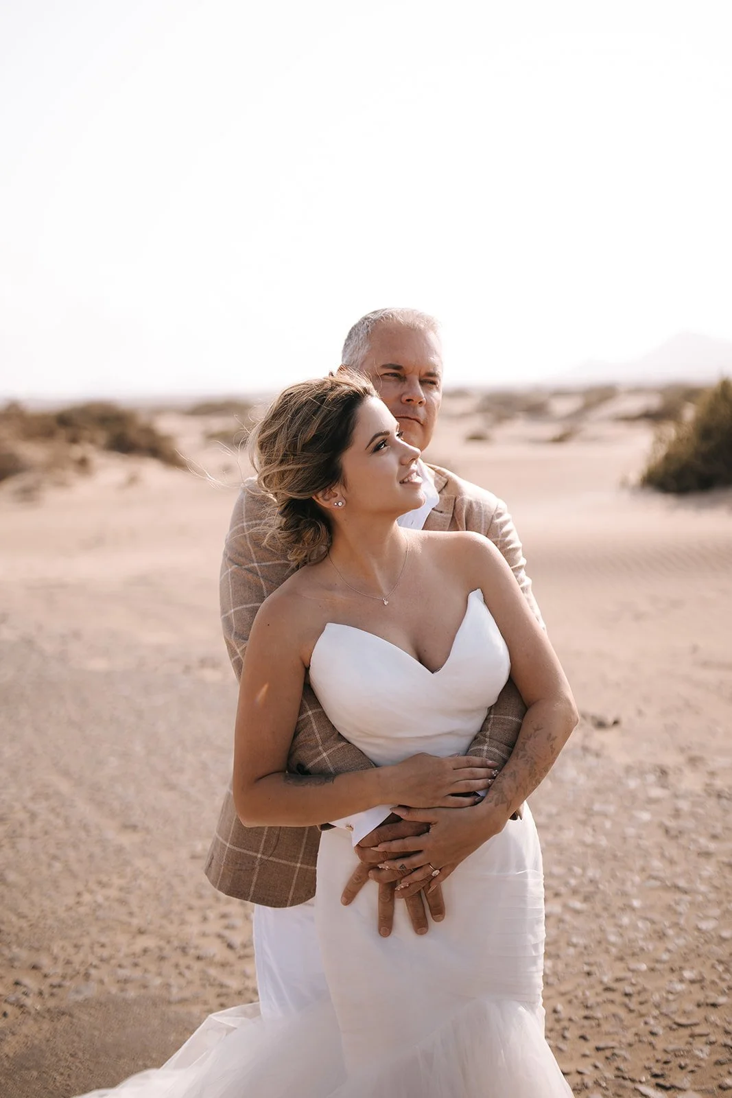 A bride and groom embrace in a desert landscape during a wedding photoshoot. The bride is in a white strapless wedding gown, and the groom in a beige checked suit jacket. The background shows sandy terrain with sparse bushes under a bright sky.