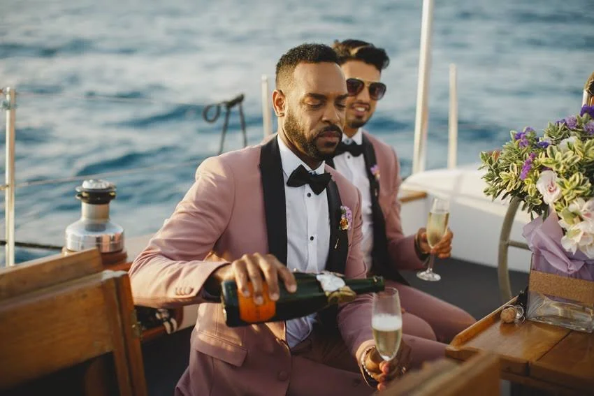 Two men in pink tuxedos with bow ties on a boat, pouring champagne and smiling, with ocean and floral decorations in the background.
