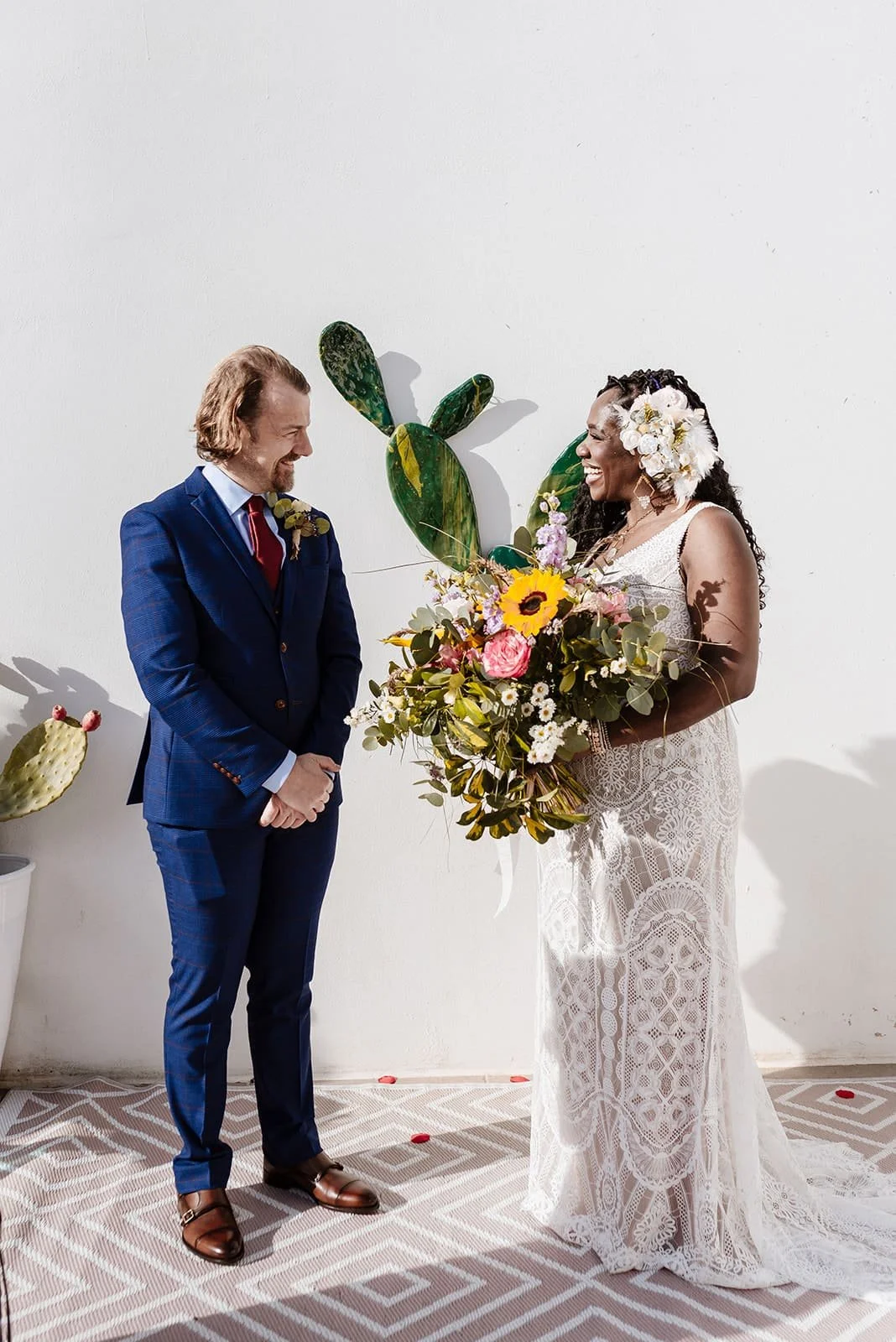A bride and groom exchanging vows during their wedding ceremony. The bride, wearing a white lace dress and floral hairpiece, holds a large bouquet of flowers including sunflowers, pink roses, and greenery. The groom is dressed in a blue suit with a r