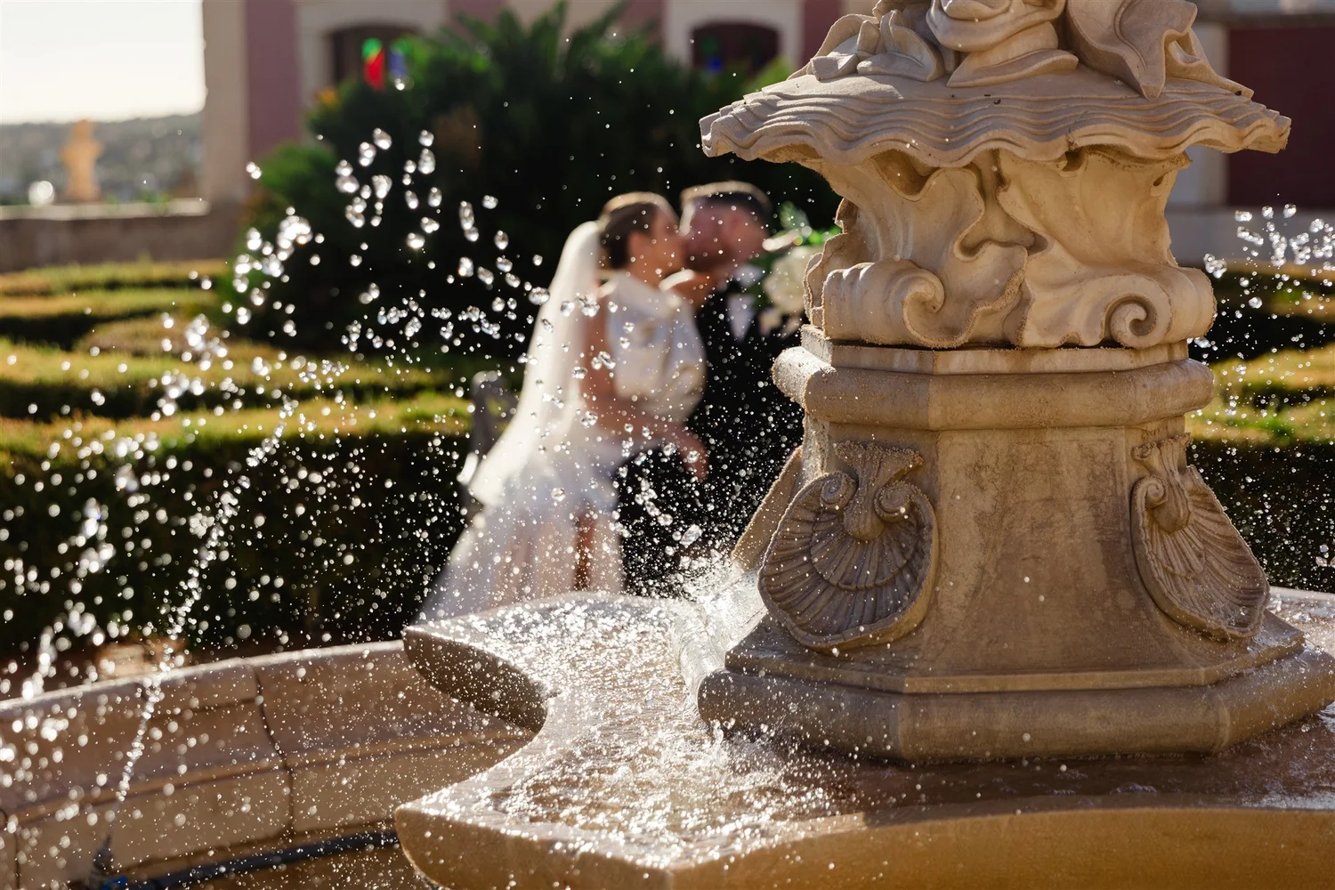 A couple, dressed in wedding attire, share a kiss behind a fountain with water spraying from its ornate stone structure.