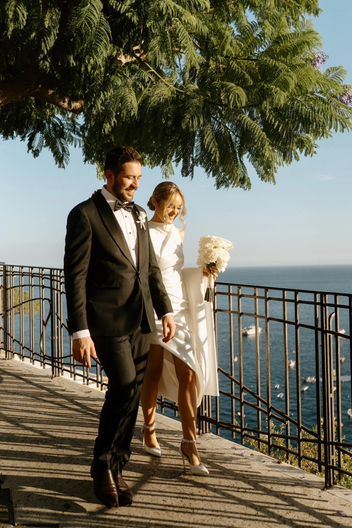 A groom in a black tuxedo and a bride in a white wedding dress walk along a seaside promenade. The bride holds a bouquet, and both are smiling. A large tree with lush green leaves shades them, and the sea with boats is visible in the background.