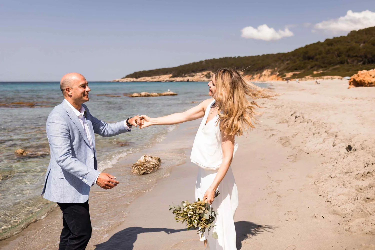 A man in a light gray blazer and a woman in a white dress exchange vows on the beach. The woman holds a bouquet and the man is holding her hand. The beach has rocks in the sand, with a rocky hillside in the background and a few people in the distance