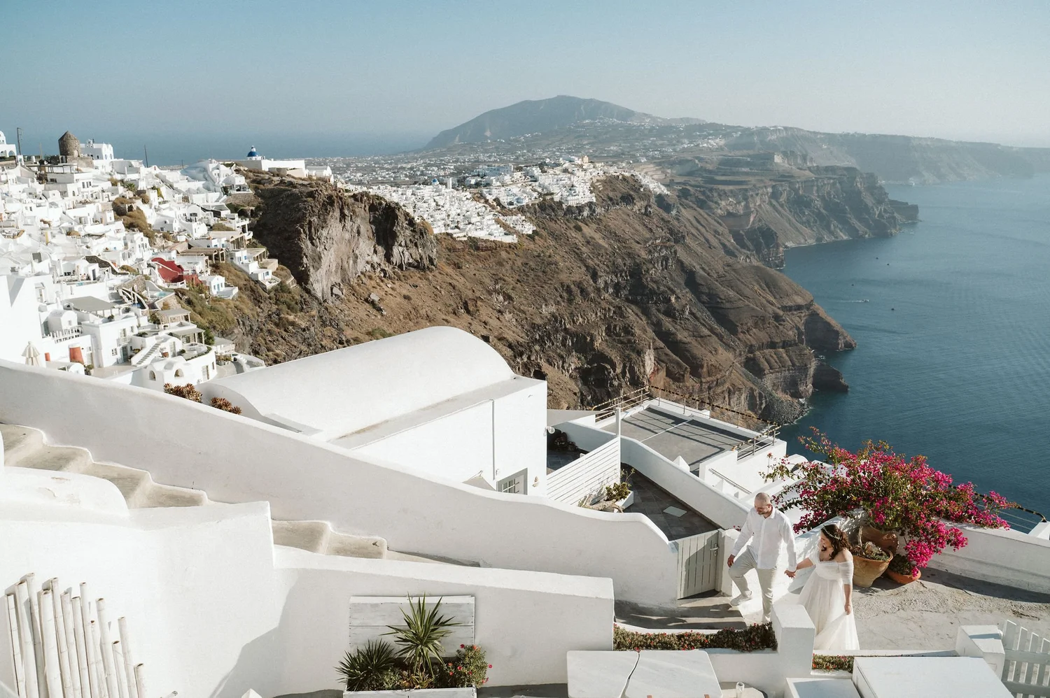 A couple dressed in white, holding hands, walking down white stairs on a hillside with white buildings, pink flowers, and the sea in the background.