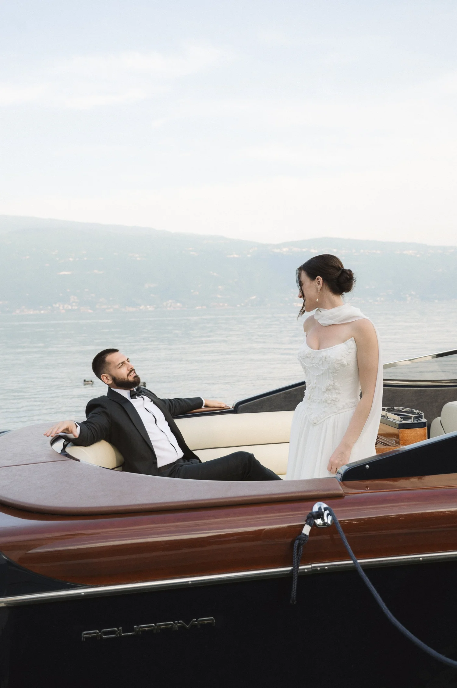 A man in a tuxedo sitting in a boat and looking up at a woman standing in front of him outdoors by a lake with mountains in the background. The woman is wearing a white dress and looking down at the man.