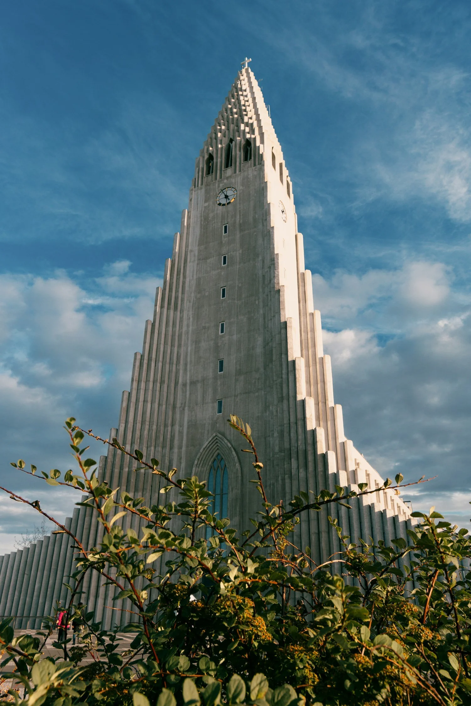 A tall, white church steeple with a cross on top, against a blue sky with scattered clouds, partially obscured by green foliage in the foreground.
