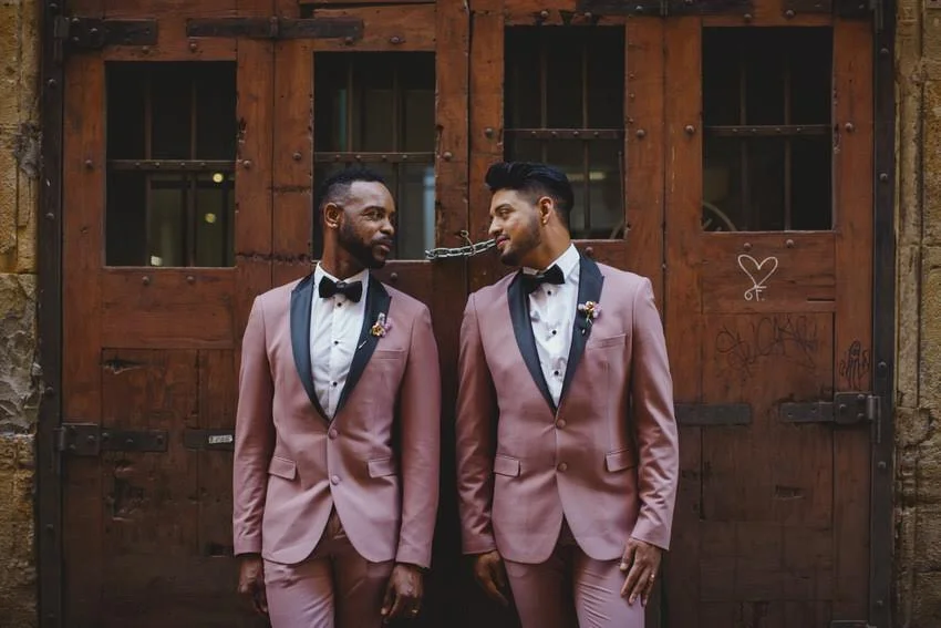 Two men in matching pink tuxedos with black lapels and bow ties standing in front of a wooden door chained together with a chain around their necks, looking at each other and smiling.