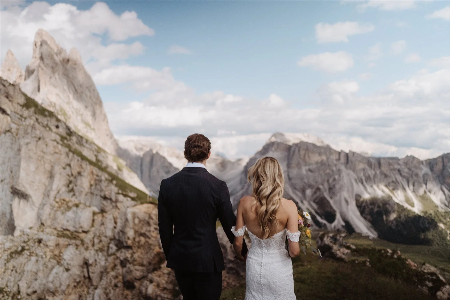 A couple, dressed in wedding attire, walking hand-in-hand through a mountainous landscape with rocky formations and cloudy sky.