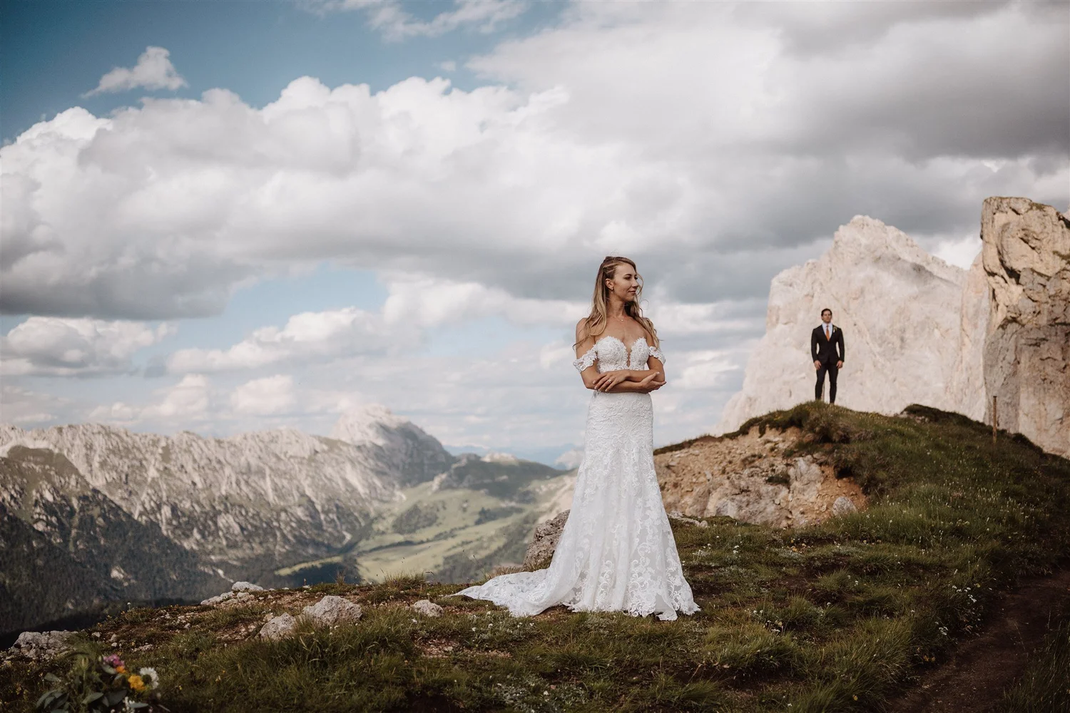 A woman in a white wedding dress stands on a mountain ledge with a scenic view of mountains and a cloudy sky in the background. A man in a suit is standing further away near a large rock.