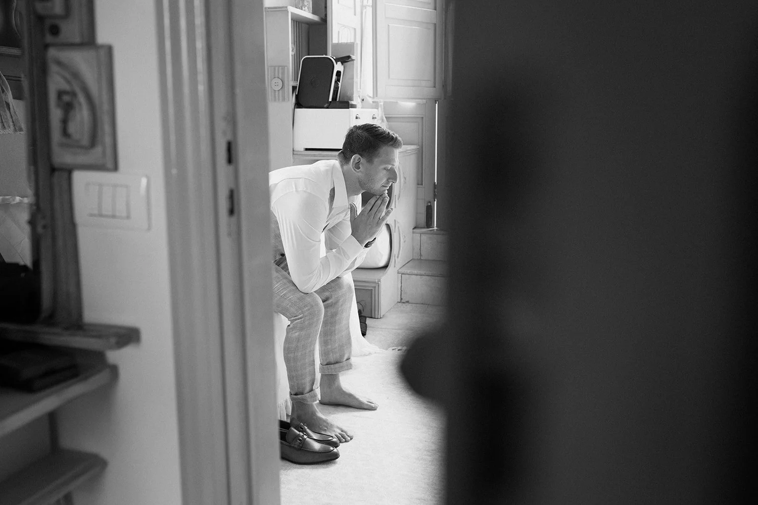 A man in a white shirt and checkered pants, barefoot, is sitting on a step with hands clasped in prayer or contemplation. He is partly obscured by a dark shape in the foreground, and appears to be in a kitchen or similar room with cabinets and a set