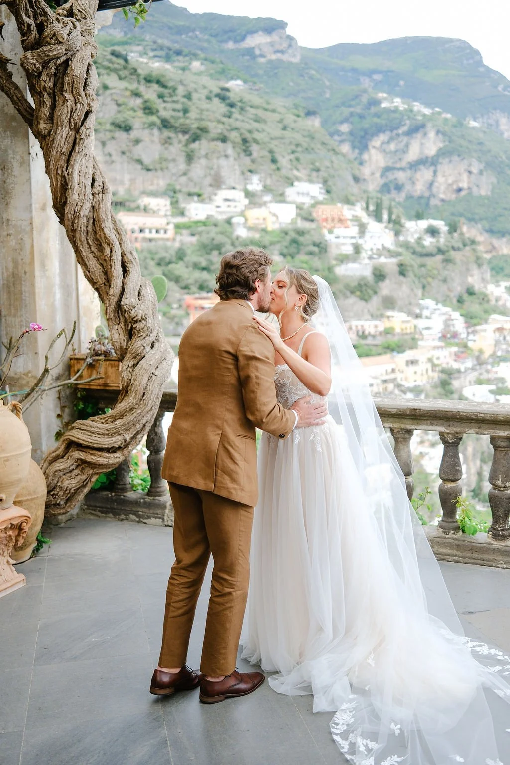 A bride and groom share a kiss on a scenic balcony overlooking a hilltop town, with the bride wearing a white wedding dress and a veil, and the groom in a brown suit.