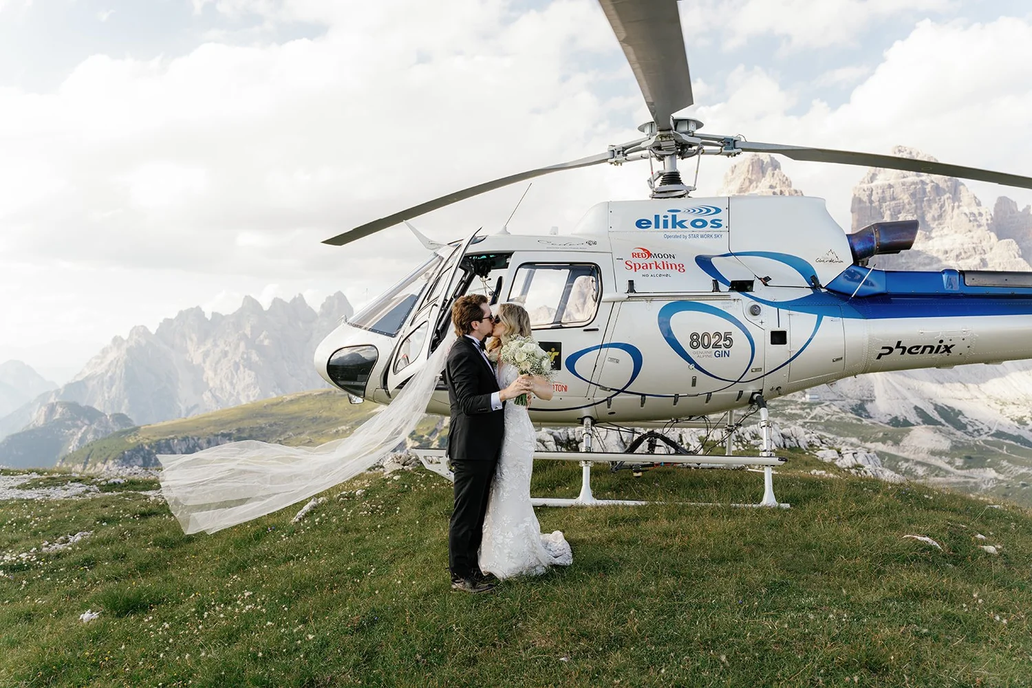 A bride and groom standing close together, kissing, in front of a helicopter on a mountain meadow. The bride is holding a bouquet and wearing a white wedding dress with a long veil. The groom is dressed in a black suit, and the backdrop features rugged mountain peaks under a partly cloudy sky.