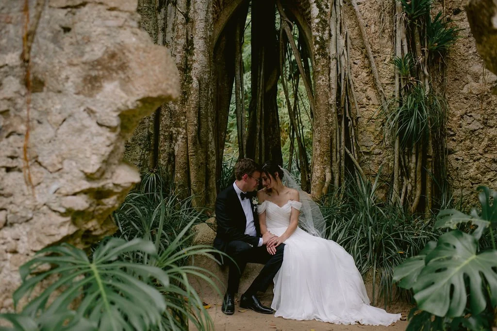 A bride and groom sitting closely together in a lush tropical garden with large roots and foliage surrounding them.