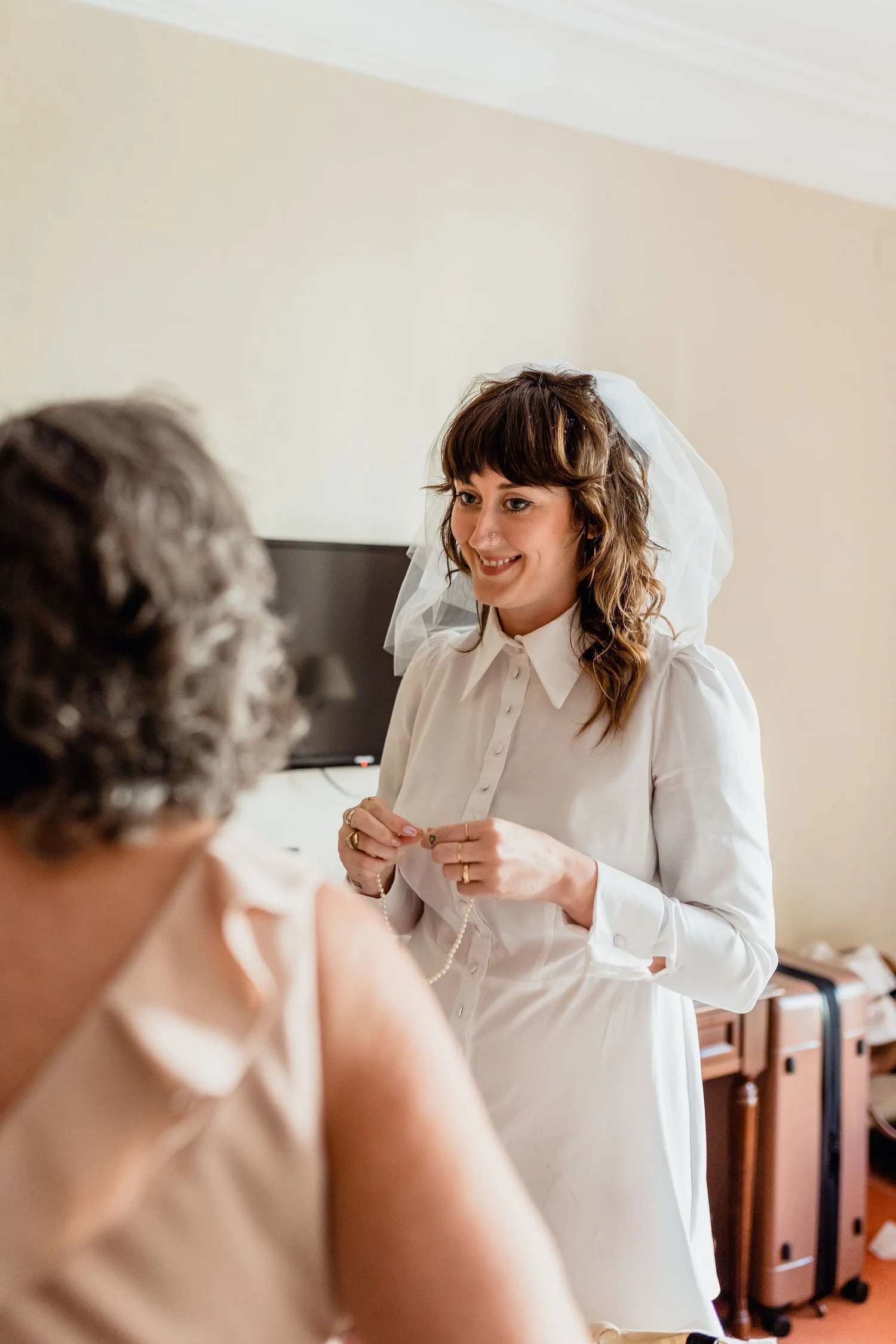 A woman in a white dress and veil, smiling, appears to be preparing for a wedding or special event, with a person in the foreground partially out of focus.