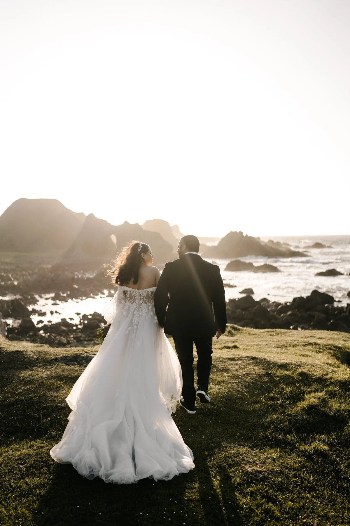 Couple dressed in wedding attire walking along a grassy path near the ocean, with rocky cliffs and the setting sun in the background.