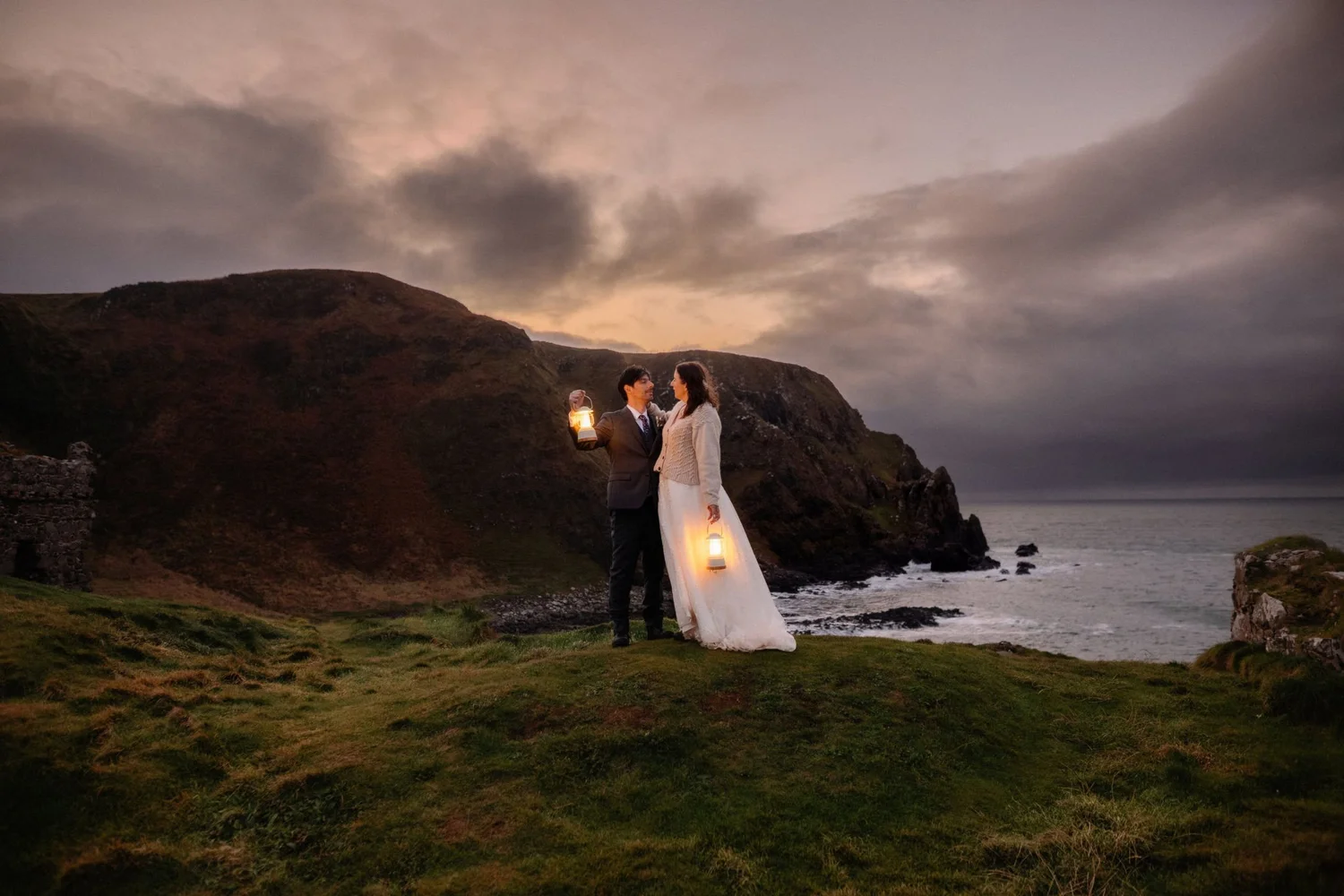 A couple dressed in formal wedding attire stands on a grassy cliffside overlooking the ocean at dusk. The man holds a lantern, and both are holding lanterns, with the sky cloudy and the landscape dimly lit.