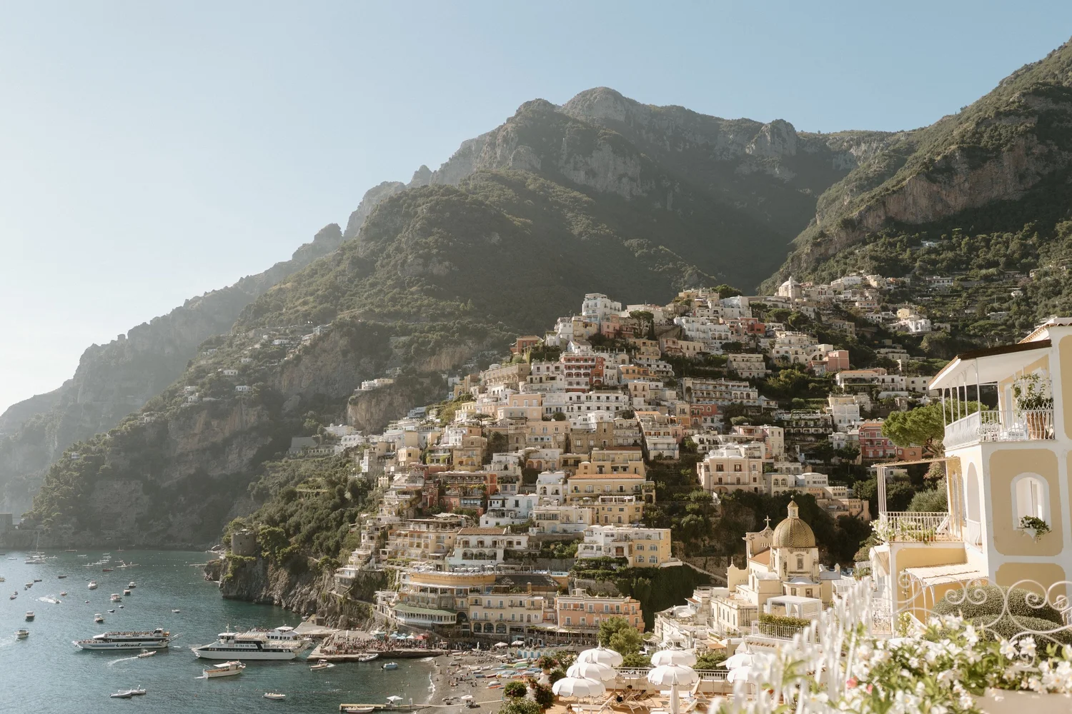 A coastal town built into steep green mountains, with colorful buildings, a sandy beach, and boats in the water.