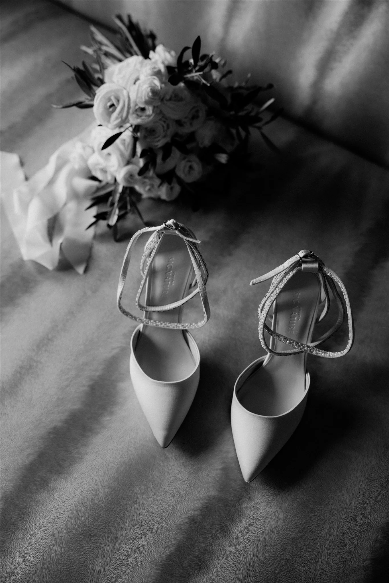 A pair of high-heeled shoes with embellished straps, placed on a textured surface. In the background, a bouquet of flowers lying on a fabric surface.