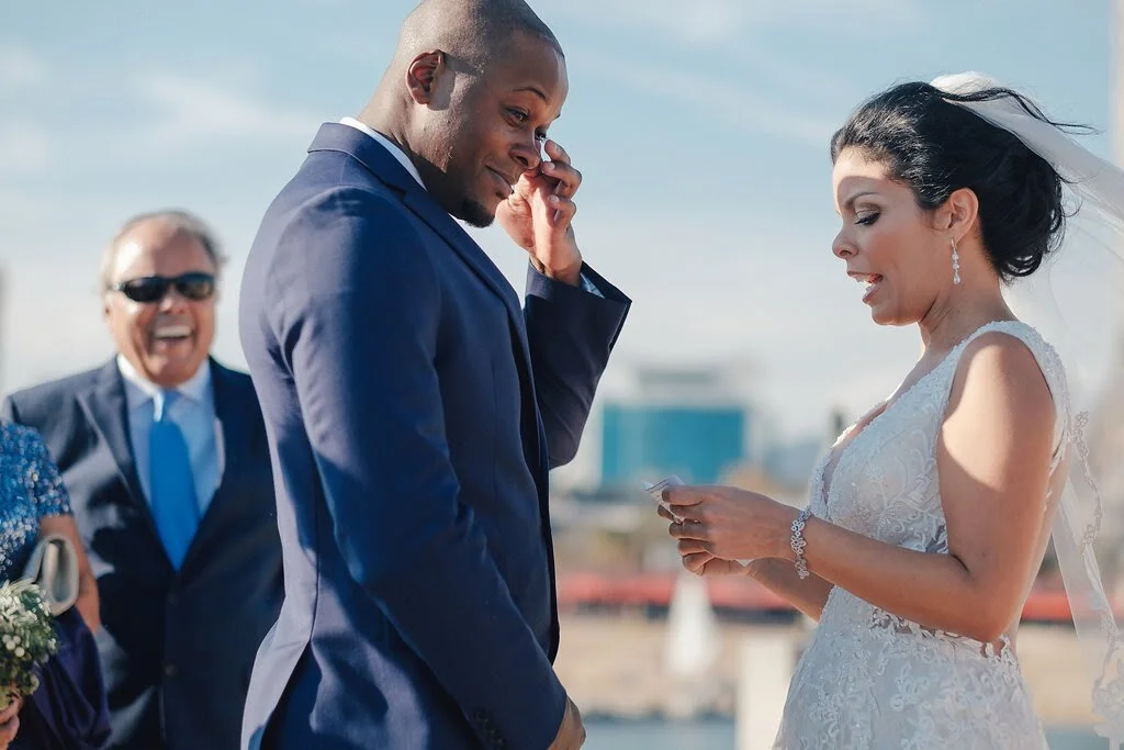 A bride and groom exchanging vows during wedding ceremony outside, with a happy older man in the background.