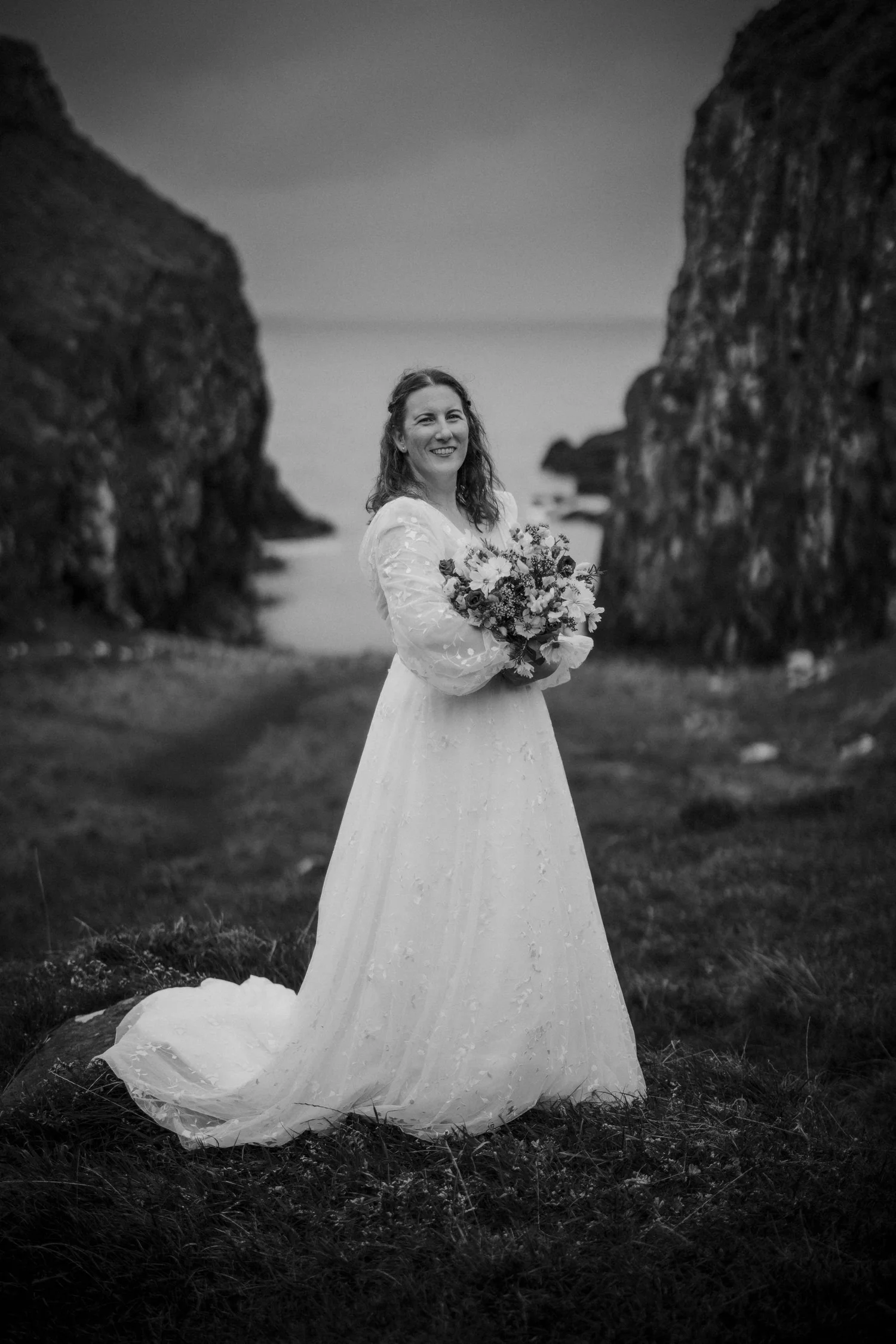 Woman in wedding dress holding a bouquet of flowers, standing outdoors between large rocks with a blurred landscape in the background.