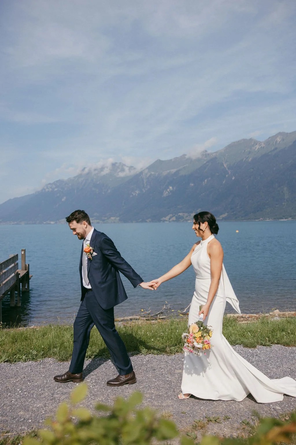 A bride and groom holding hands and walking near a lake with mountains in the background. The groom is wearing a navy suit and the bride is in a white wedding dress, holding a bouquet of flowers.