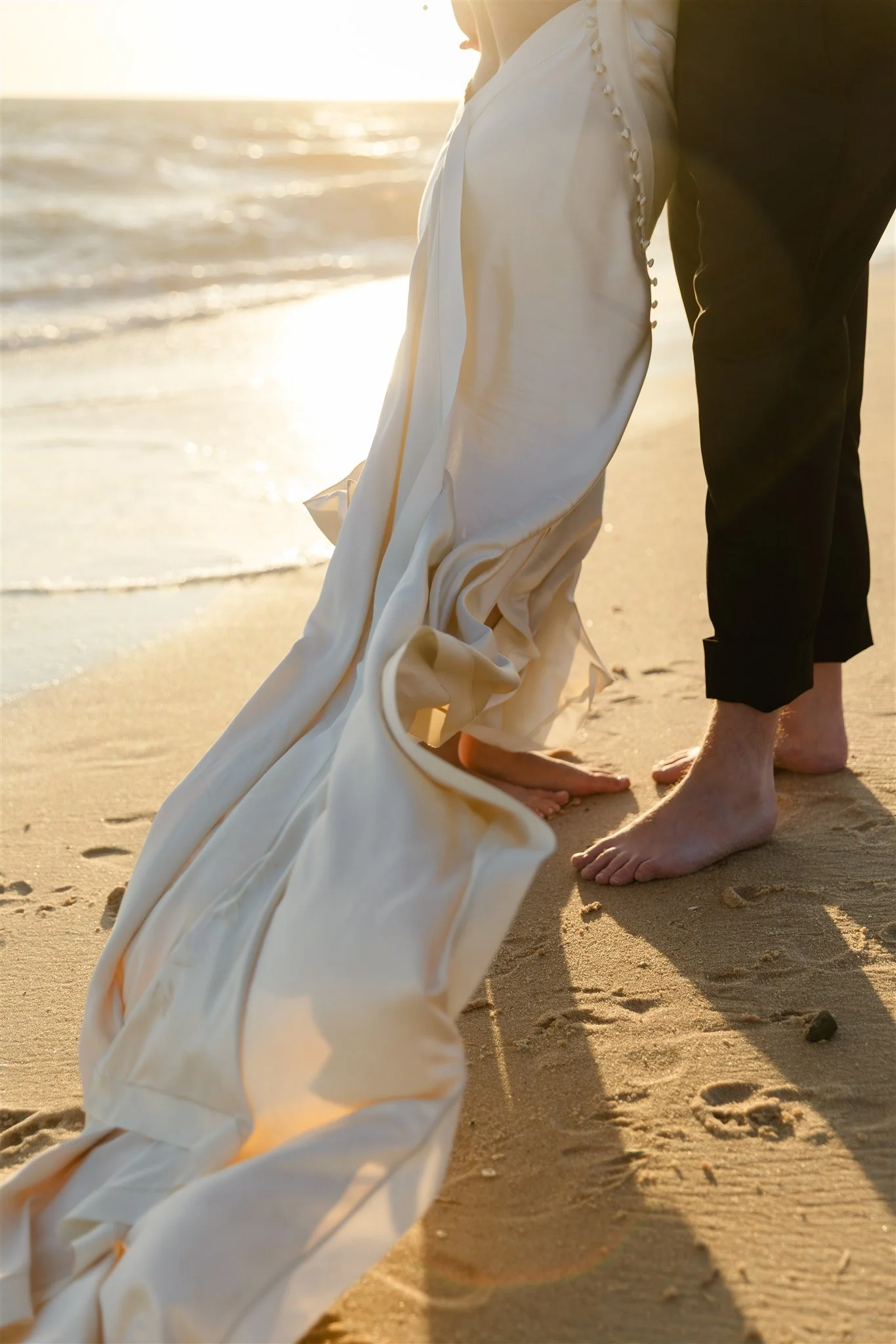 Close-up of a couple standing barefoot on a sandy beach during sunset, with the woman wearing a white wedding dress and the man in black trousers, holding hands.