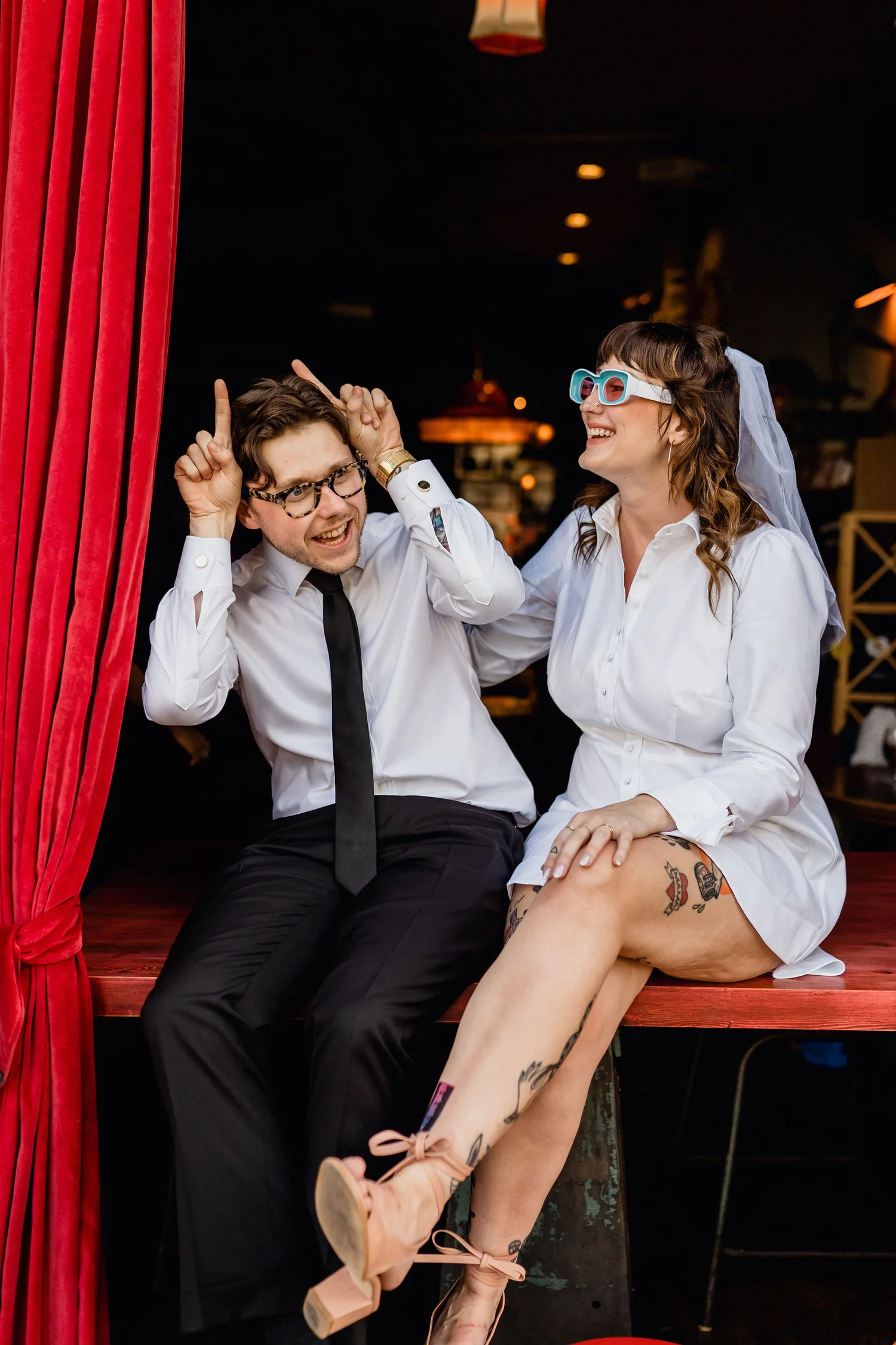 A man and a woman sitting on a wooden bench behind a red curtain in a dimly lit room, enjoying a playful moment. The man is wearing a white shirt, black tie, and glasses, with his hands up near his head, holding his ears. The woman is wearing a white