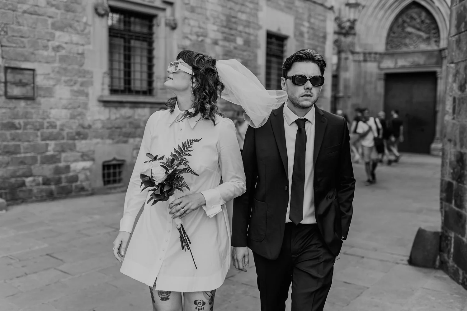 A black-and-white photo of a bride and groom walking outdoors near a stone building. The bride wears a white dress, glasses, and a veil, and holds a bouquet of flowers. The groom wears a dark suit, tie, and sunglasses. They walk closely, with the bri