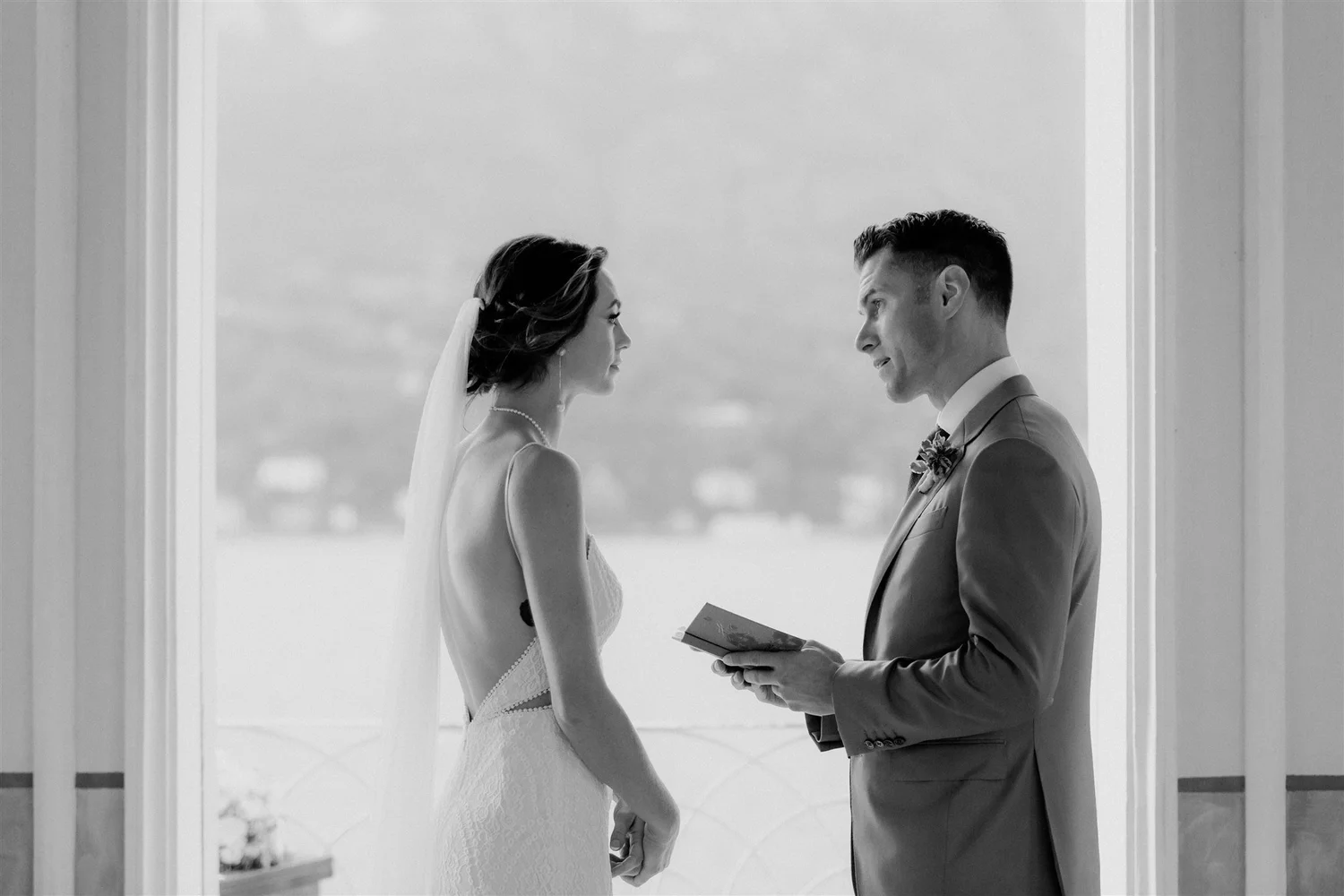 A bride and groom standing face to face in a doorway, holding hands, preparing for their wedding ceremony. The bride is in a white lace wedding dress with a veil, and the groom is in a light-colored suit with a tie and boutonniere.