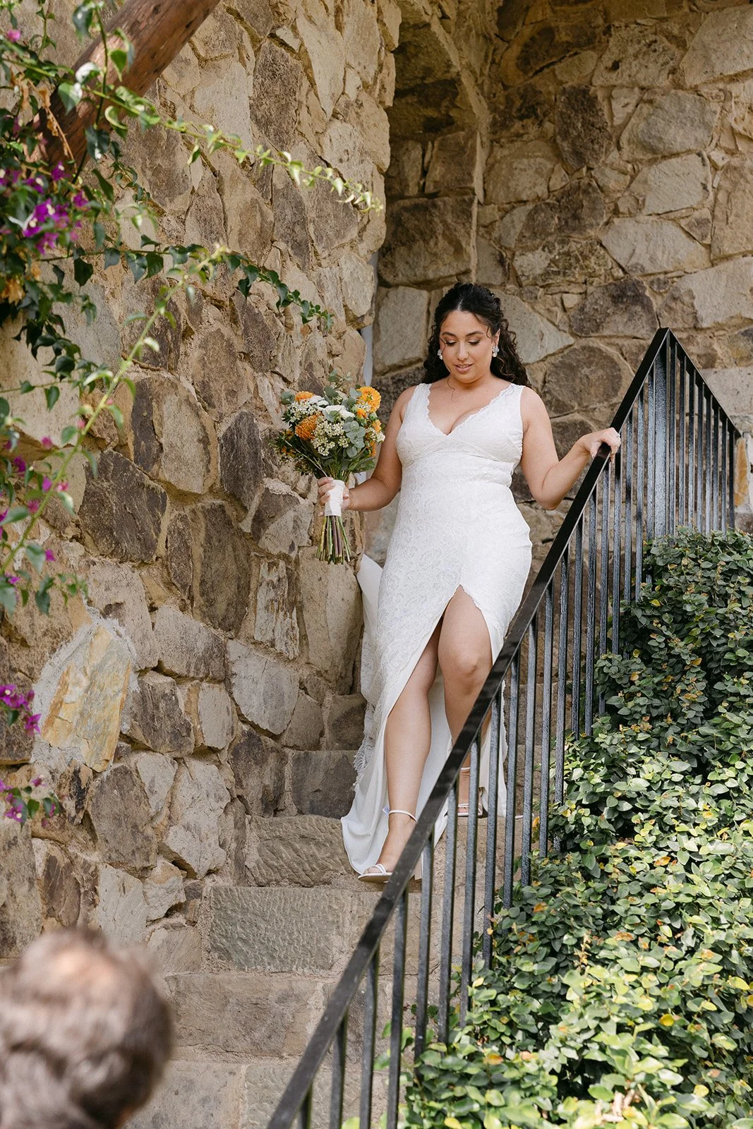 Bride in a white lace dress with a high slit, holding a bouquet of flowers, walking down stone steps beside a stone wall and black metal railing, surrounded by greenery.
