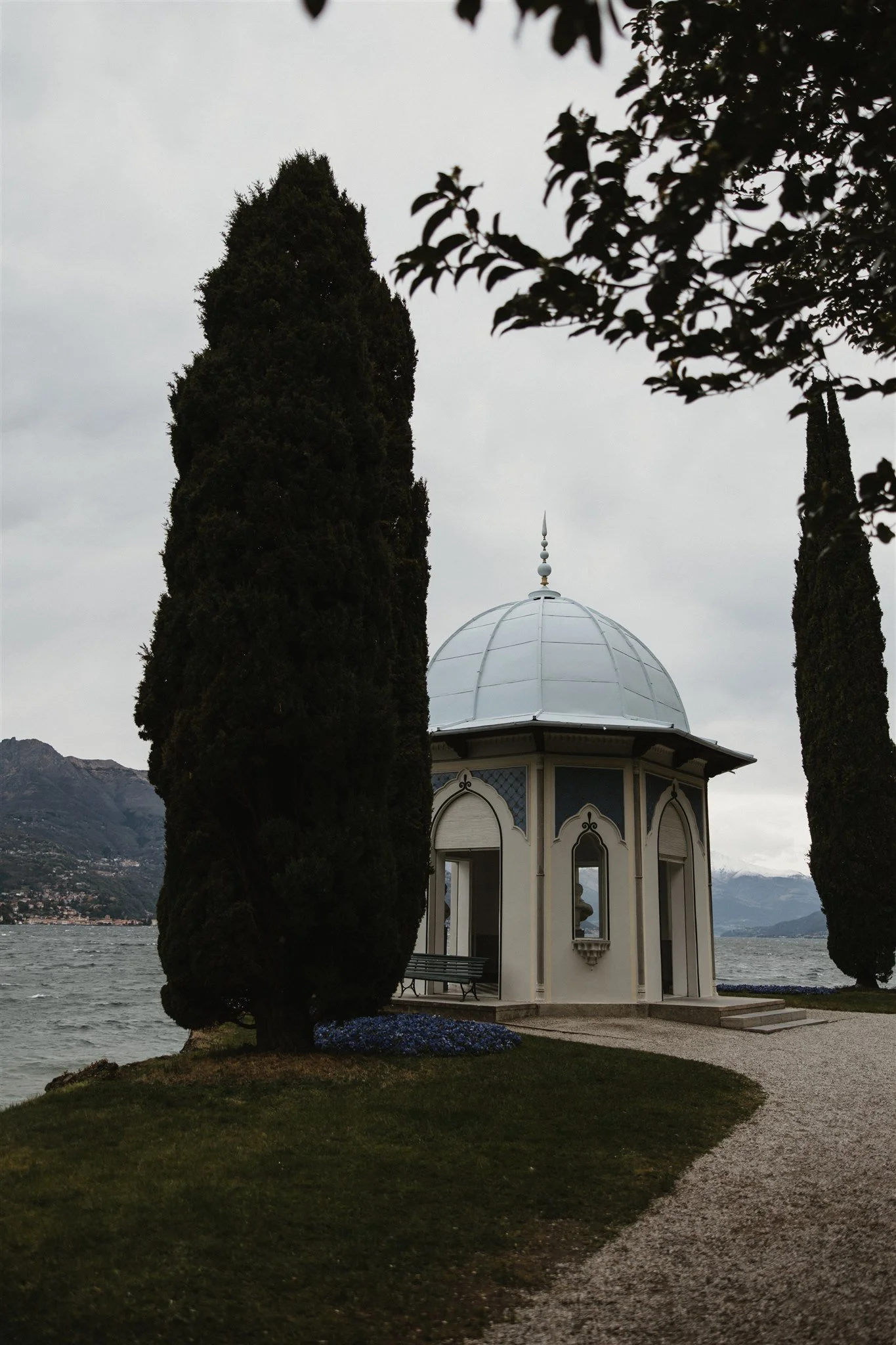 A lakeside pavilion with a domed roof, surrounded by tall cypress trees, with mountains and water in the background on a cloudy day.