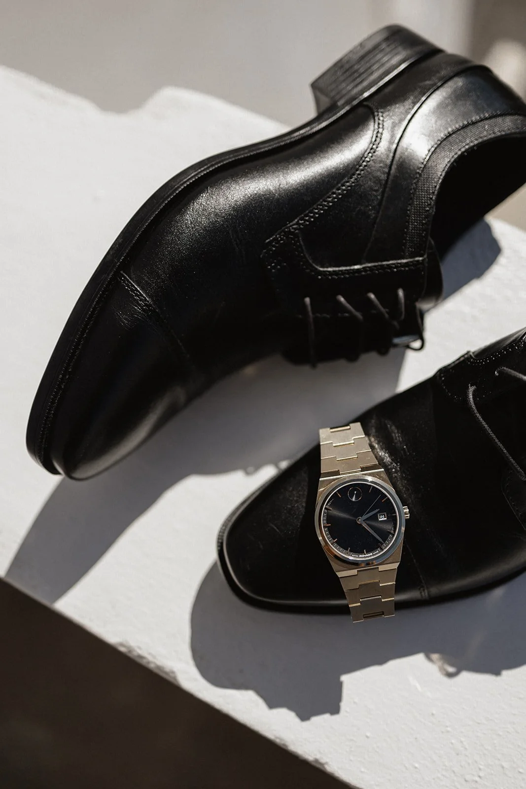 A black leather dress shoe and a silver wristwatch with a black face, resting inside an open black leather shoe box, with shadows cast on a white surface.