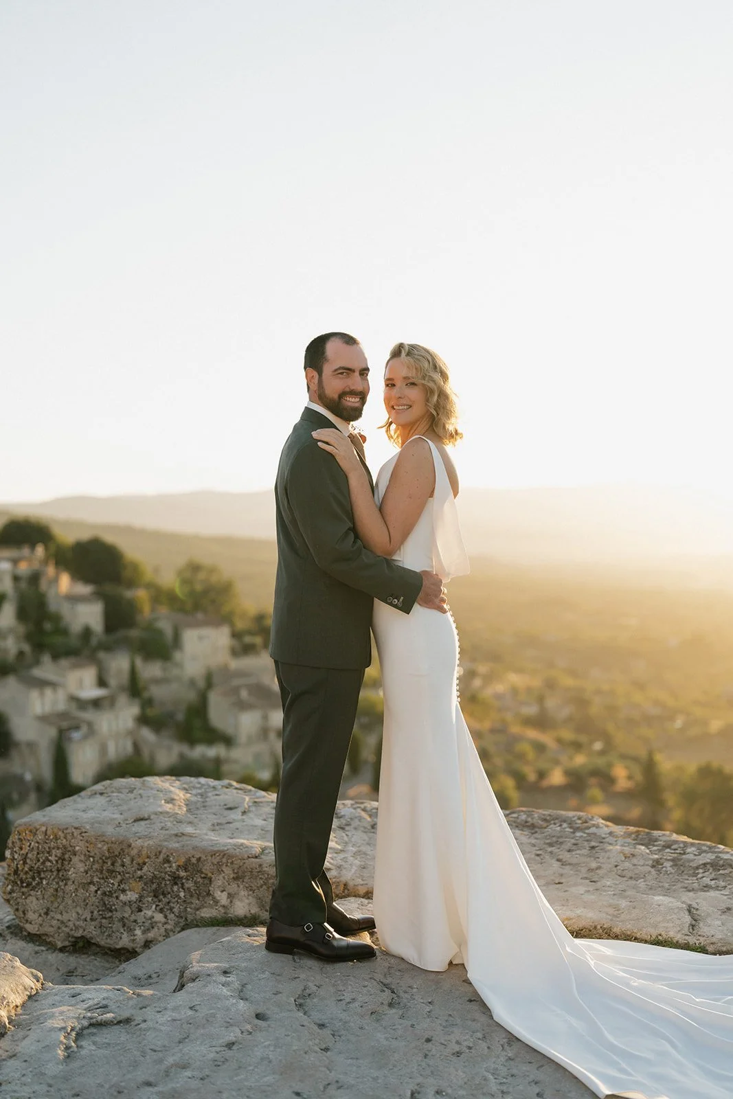 A newlywed couple stands on a rocky ledge outdoors during sunset, with a scenic landscape of houses and rolling hills in the background, dressed in wedding attire, smiling and holding each other.