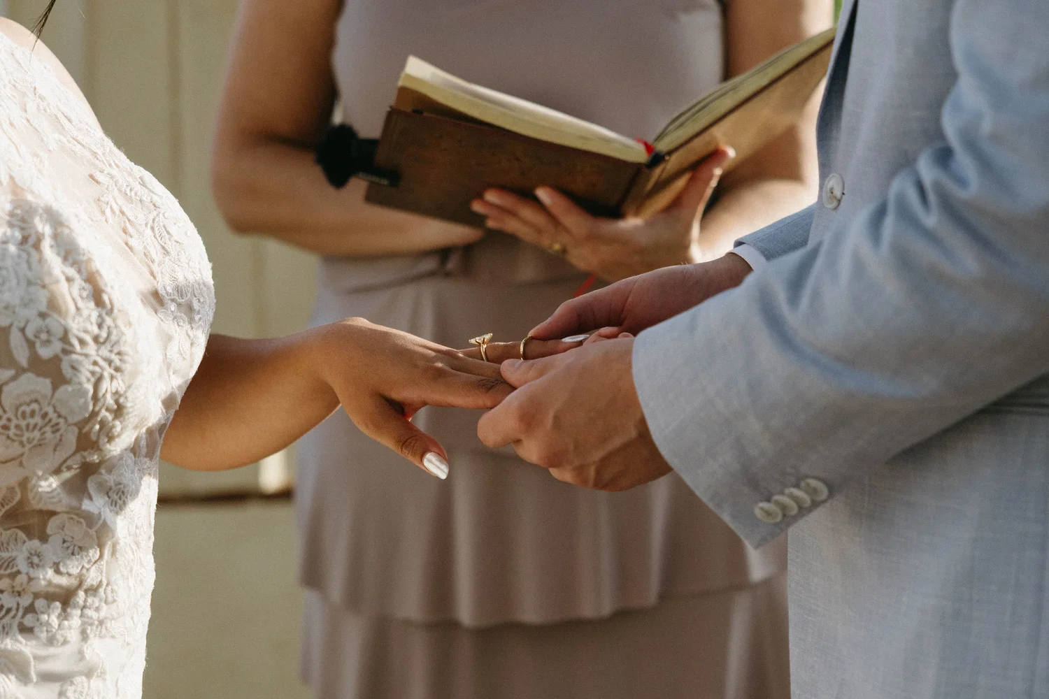 A close-up of a wedding ceremony, showing the couple holding hands and exchanging rings, with an officiant present in the background holding a book.