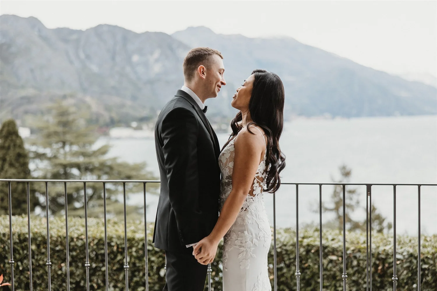 A bride and groom stand close together, holding hands, looking at each other, with a scenic mountain and lake background.