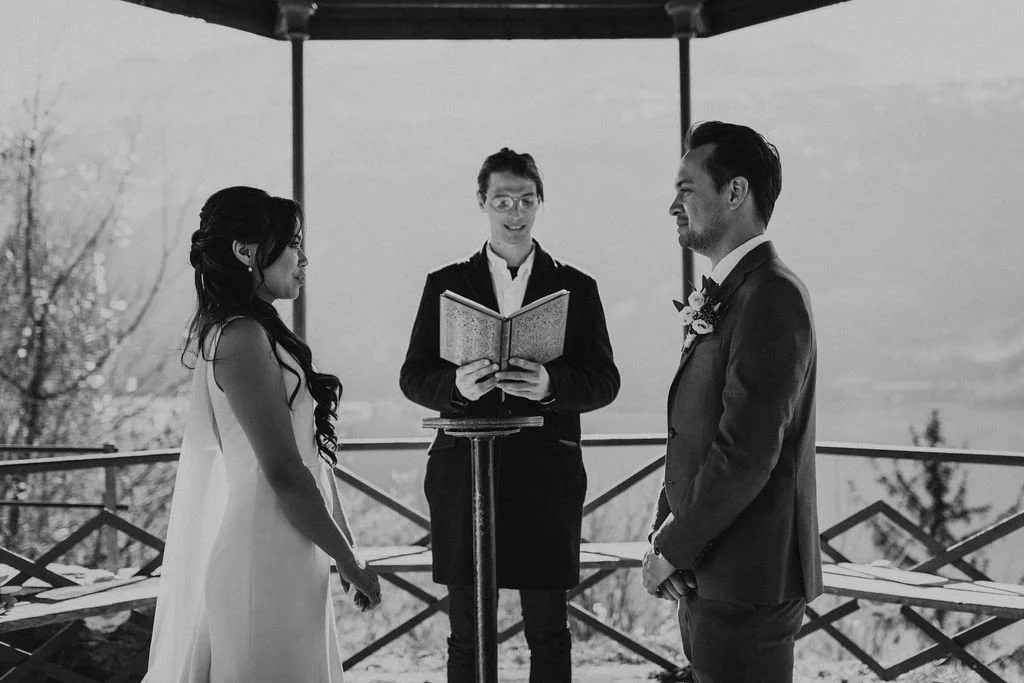 A black and white photo of a wedding ceremony with a bride and groom standing face to face under a canopy, with an officiant in the middle reading from a book.