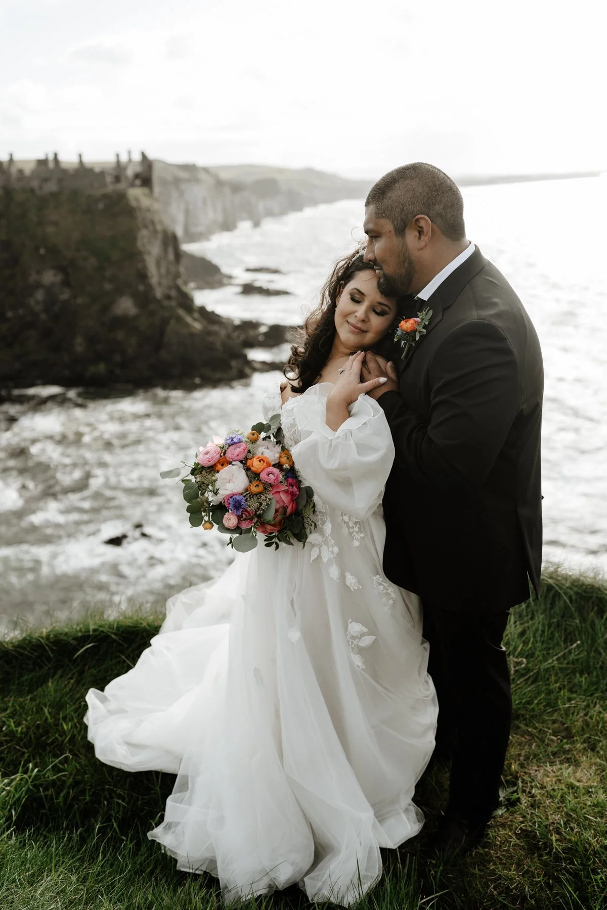 A bride and groom embrace by the ocean on a grassy patch. The bride is holding a colorful bouquet and wearing a white wedding dress, while the groom is dressed in a black suit with a boutonniere. In the background, cliffs and the sea are visible.