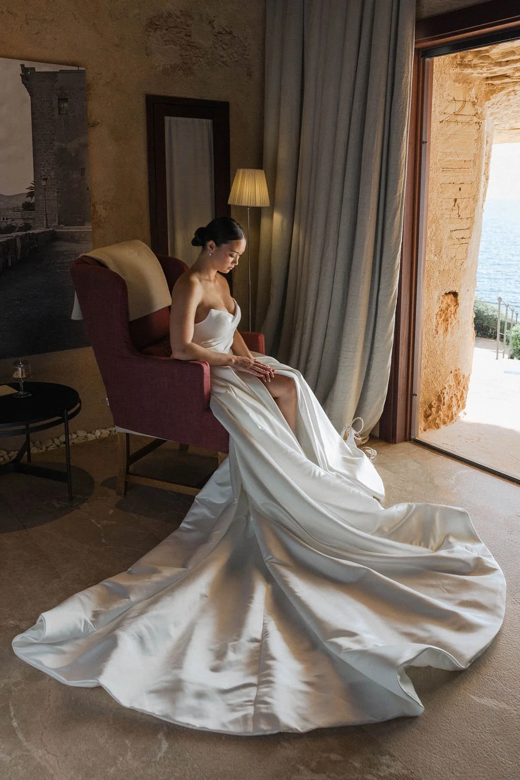 A woman in a white wedding dress sitting on a red armchair by a large open window overlooking the ocean.
