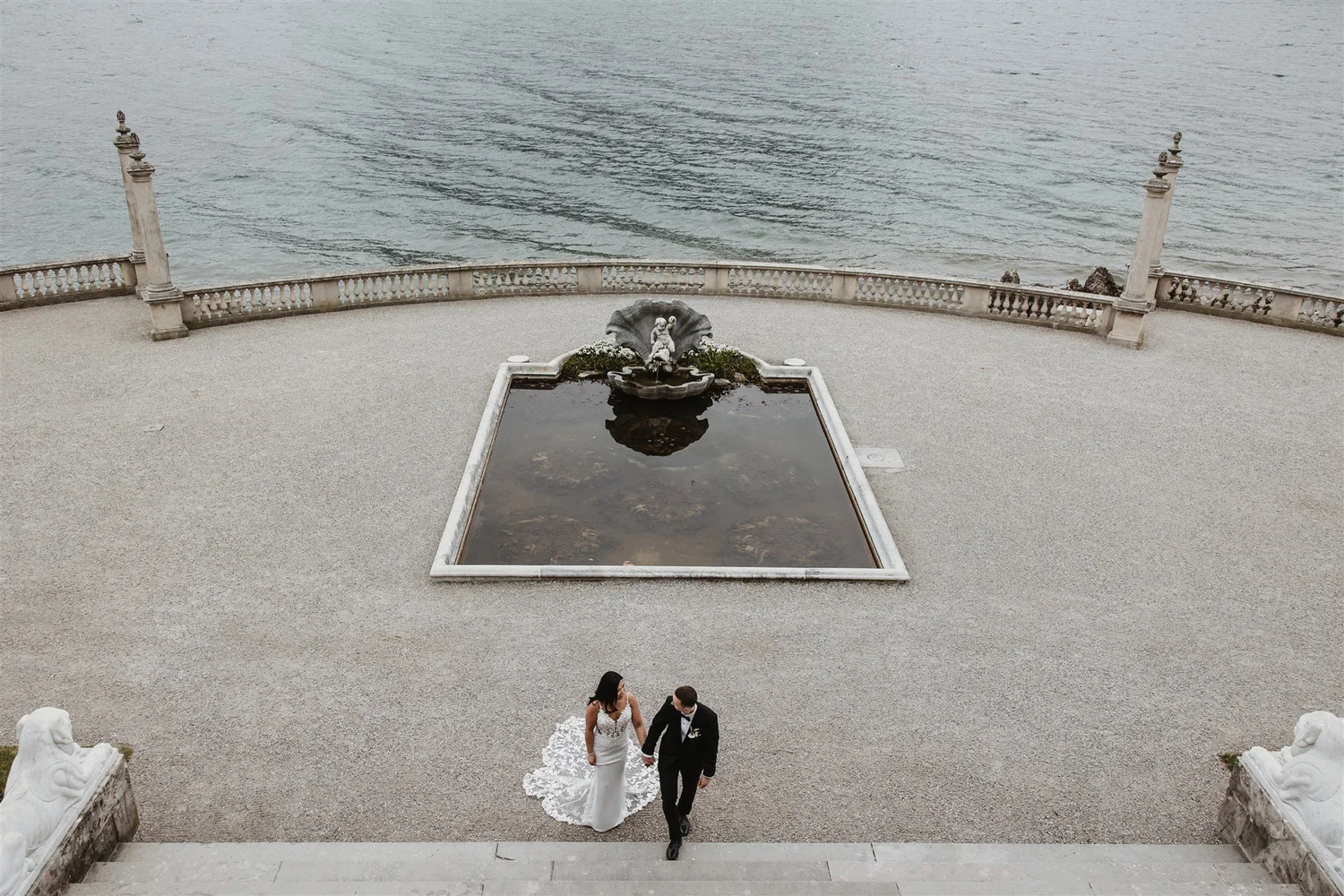 A wedding couple holding hands and walking down a stone staircase toward the seafront, with a stone fountain nearby, large water body in the background, and a decorative stone balustrade