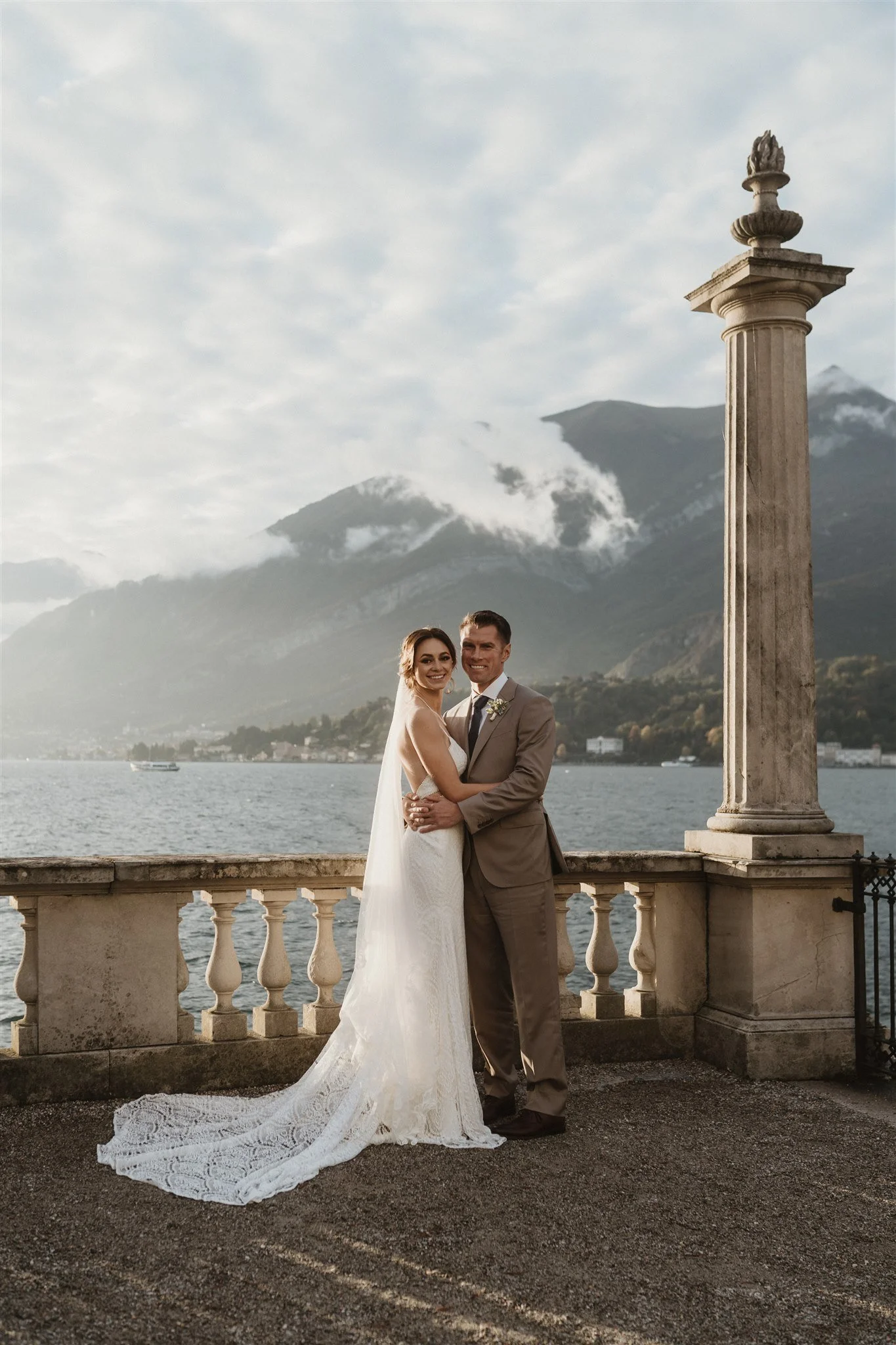 A bride and groom standing close together on a balcony overlooking a body of water, with mountains in the background. The bride is in a white lace wedding gown with a long train, and the groom is in a tan suit. There is a tall stone column with a dec