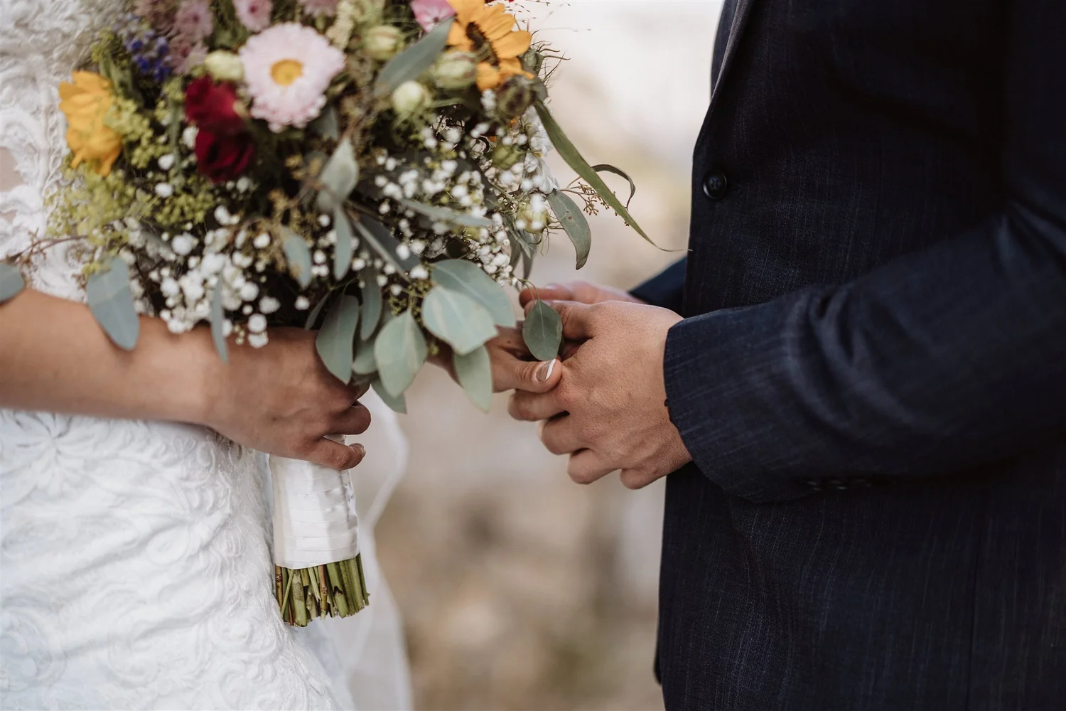 A woman in a white lace dress holding a bouquet of flowers, standing close to a man in a dark suit, during a wedding ceremony.