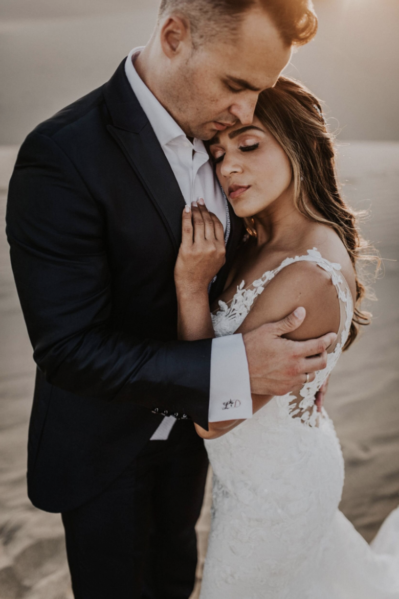 A newlywed couple embracing on a beach during sunset, with the groom in a black tuxedo and the bride in a white lace wedding dress.